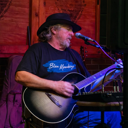 Man Johnny Winkle performing live music wearing a black BleuMondaze Live Stream Podcast t shirt with blue brushstroke logo while playing acoustic guitar on stage