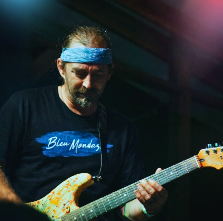 Guitarist wearing a black BleuMondaze Live Stream Podcast t shirt with blue brushstroke logo and blue bandana while performing on stage