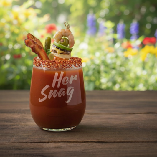 Her Snag" 20.5oz etched stemless wine glass styled as a Bloody Mary with a tajin rim and decorative garnishes on a wooden table with a garden background.