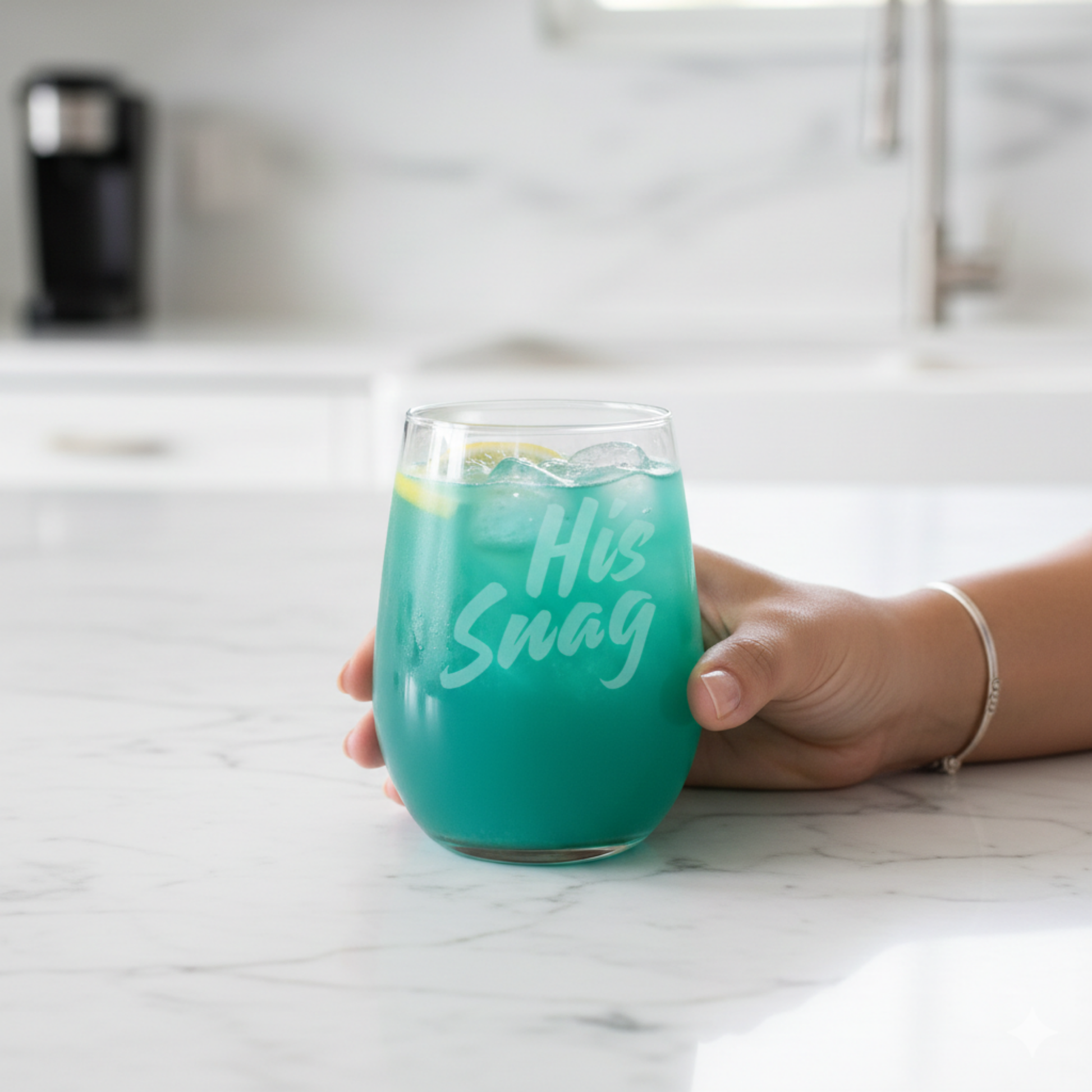 Gemini said
Hand holding a 20.5oz "His Snag" etched stemless wine glass by Expressive DeZien, filled with a teal-colored drink, on a white marble kitchen countertop.